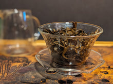 A glass gaiwan filled with glistening wet dark brown tea leaves. There is a glass pouring vessel, blurred, in the background, and they are both on a bamboo tea tray. The background is dark, blurred, and indistinct.