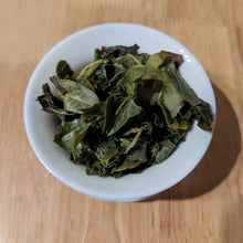 Brewed Huang Dan tea in a small white gaiwan on a wooden surface. The tea leaves are a deep bright green. Some are whole, and some are torn.