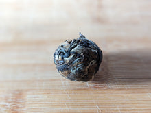 An enlarged photo of a tightly pressed ball of tea. There are varying different color shades and degrees of darkening in the tea ball. The tea ball is photographed on top of a clean bamboo cutting board.