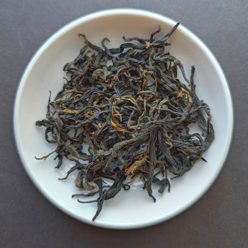Dried loose-leaf tea leaves, predominantly dark brown with subtle orange-gold tips, piled in a small white ceramic dish against a dark gray background.