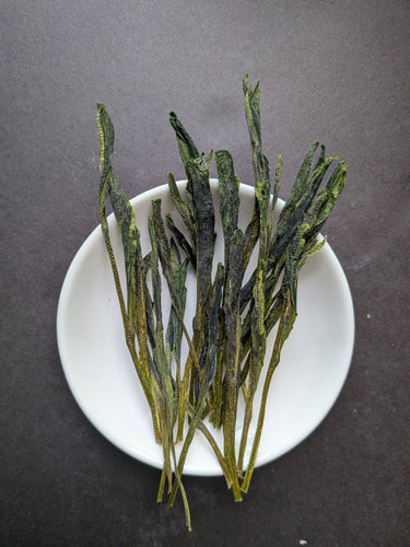 Long, flattened, green tea leaves on a small white dish against a dark grey background