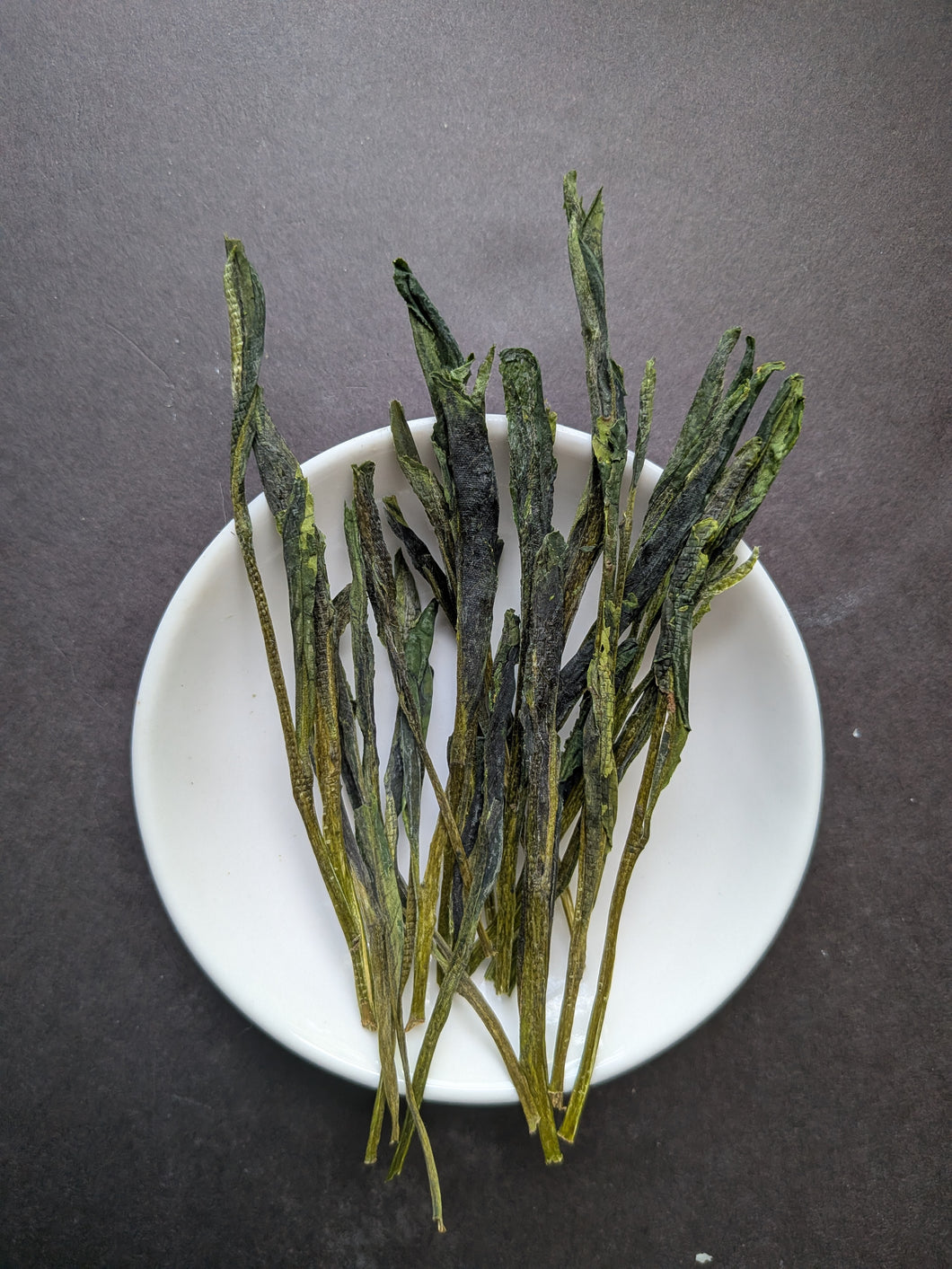 Long, flattened, green tea leaves on a small white dish against a dark grey background